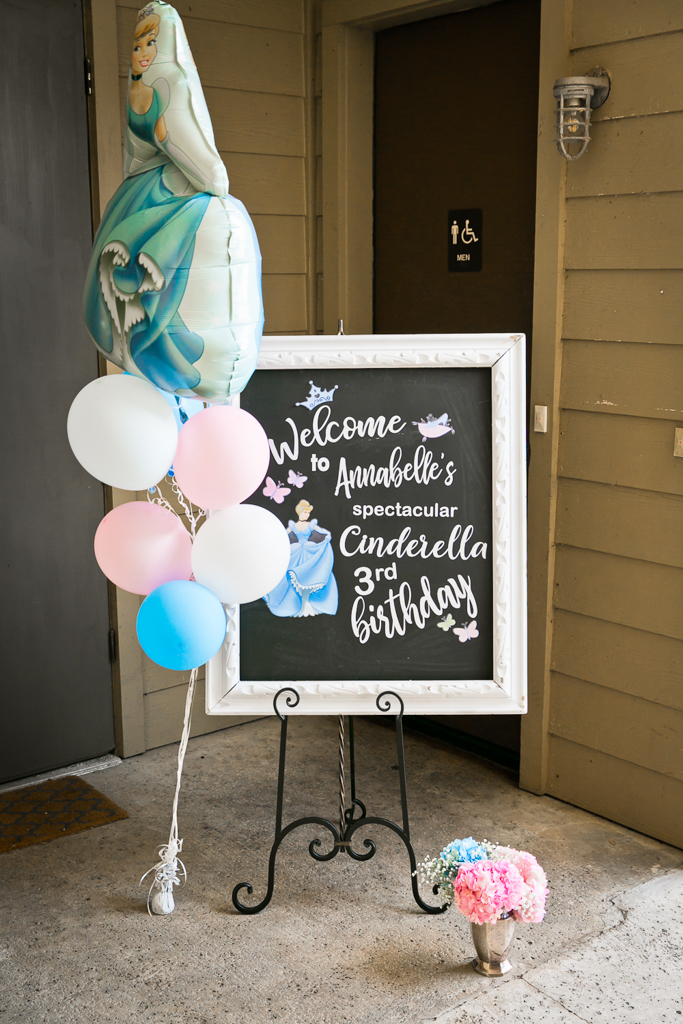 Chalkboard that says "Welcome to Annabelle's spectacular Cinderella 3rd Birthday" with pink, white and blue balloons