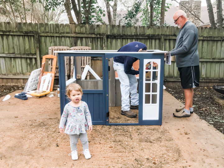Annabelle posing in front of the halfway assembled playhouse while Andrew and dad work on assembling it.
